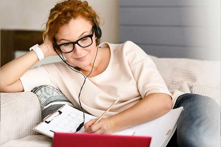 Image shows a red headed woman lounging comfortably on a couch as she takes notes and looks at an open laptop.